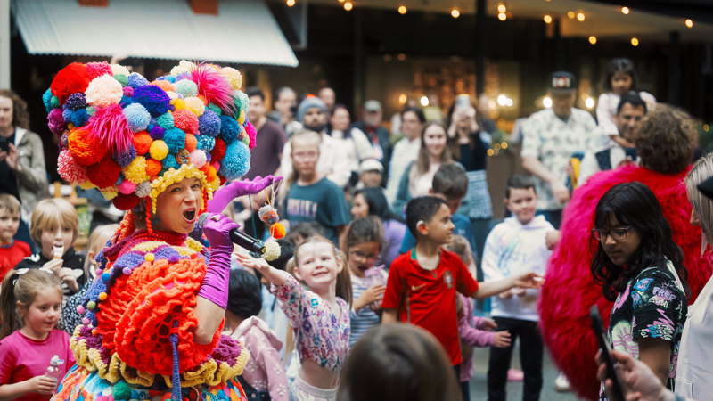 A performer holding a microphone and wearing a multi-coloured costume performs to children.