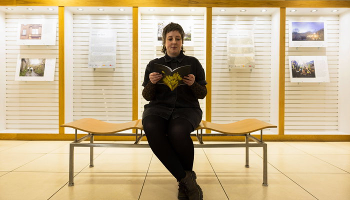 Communities and Libraries Officer Stef sits on a bench in the Mitchell Library in front of a new exhibition. She is reading a pamphlet offering tips on activities to help develop a healthier winter routine and tackle Seasonal Affective Disorder this winter. 