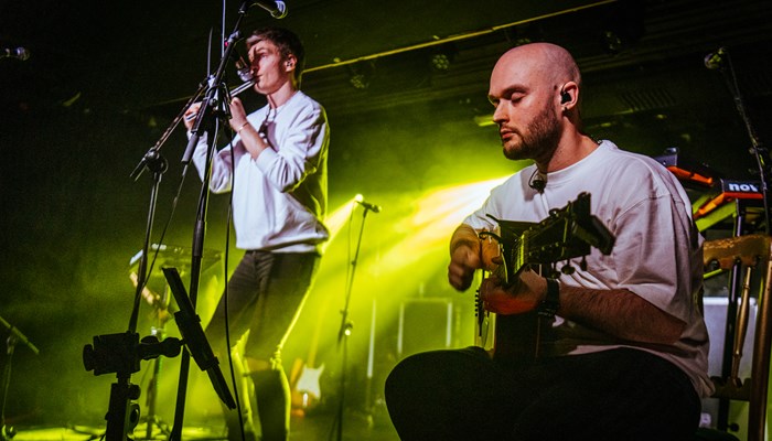 Two people on stage. One is standing wearing a light grey jumper playing a penny whistle and the other is seated wearing a white tshirt playing the guitar.