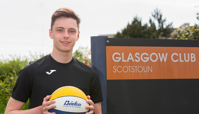 Person in black top stands holding yellow, blue and white ball in front of a sign. The sign reads 'GLASGOW CLUB SCOTSTOUN'.