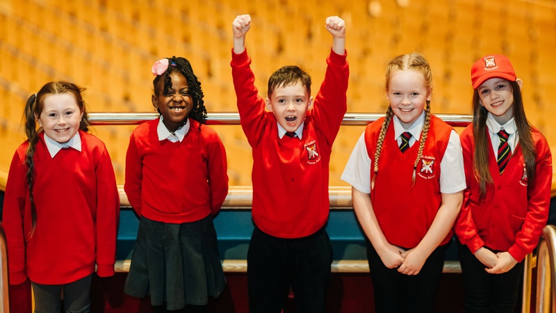 Five children smiling on the balcony of the Glasgow Royal Concert Hall, they are all wearing red school jumpers and one kid in the middle has both hands in the air.