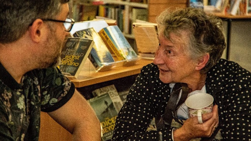 A adult and an elderly person sit in a  public library chatting. 