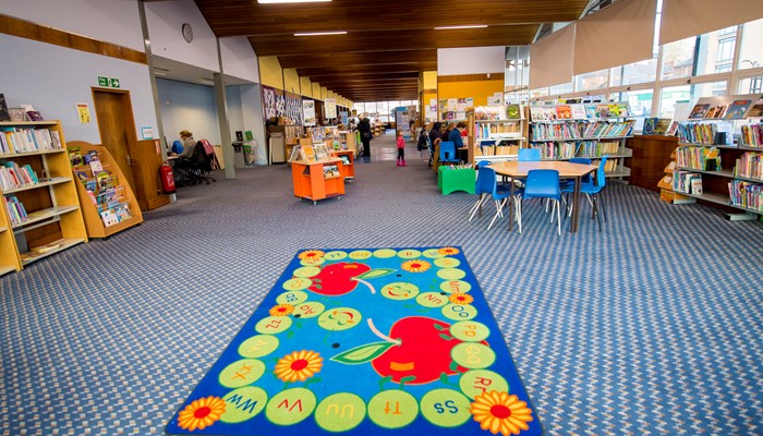 A bright, spacious children's area with a blue checked carpet, large windows along the right hand side and a wood pannelled ceiling. There is a round wooden table with blue chairs and a colourful children's rug in the centre of the room.