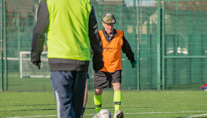 An elderly man wearing a flat cap and orange vest is walking towards a football on a green outdoor pitch.