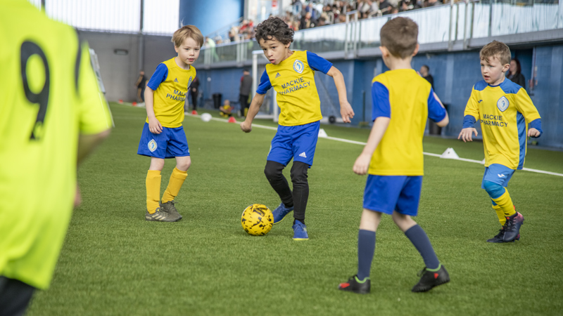 A few primary aged children play football on an indoor pitch. Each is wearing yellow and blue uniforms.
