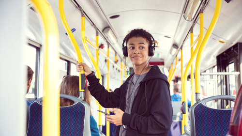 A young person wearing headphones and holding a phone is standing and smiling on a bus