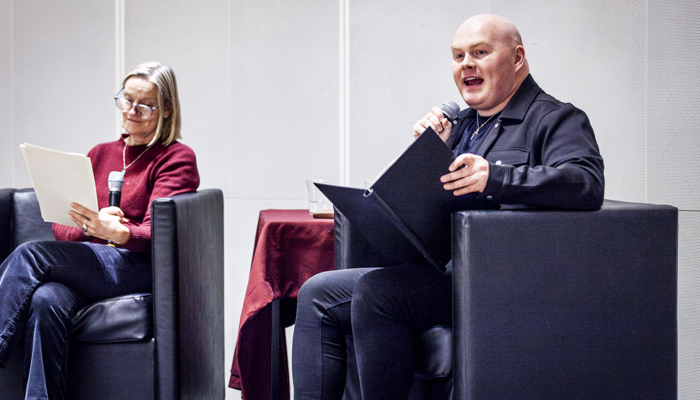 A bald man speaks with a microphone and book, next to a woman reading.