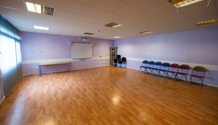 large hall with windows on the left hand side. there are 7 chairs lined up to the right of the room. the walls are painted purple and there is a table and a board from presentations at the top of the room
