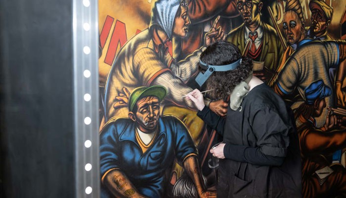 Conservator works on a detailed mural painting, using tools under bright strip lighting.