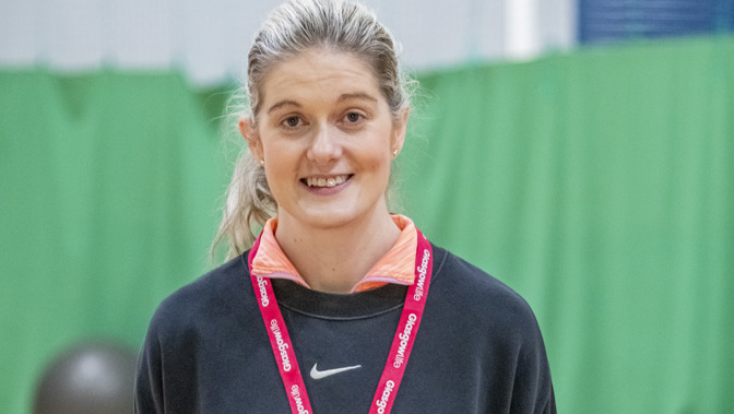A Glasgow Life employee stands indoors in a sports hall, smiling at the camera. They are wearing a dark sweatshirt over an orange top and black leggings, with a lanyard and ID badge around their neck. Their hair is tied back, and gym equipment and green wall padding are visible in the background.