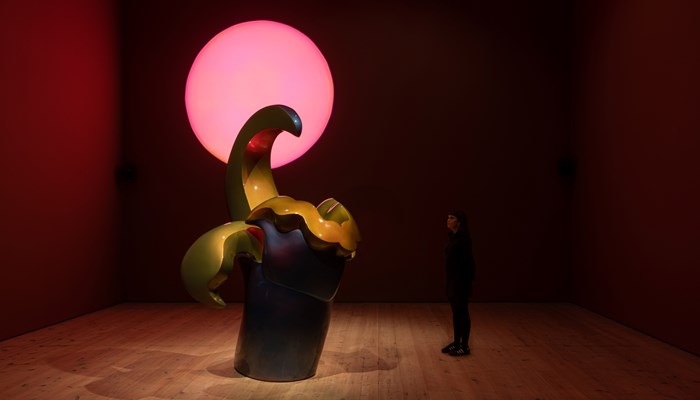 In a darkened gallery, a large abstract sculpture dwarves a lone viewer. Above them, glows a pink sun-like orb