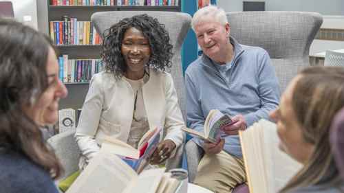 A smiling black lady and smiling white man sit in beauitful grey chairs in a library, with books in their hand, enjoying a conversation