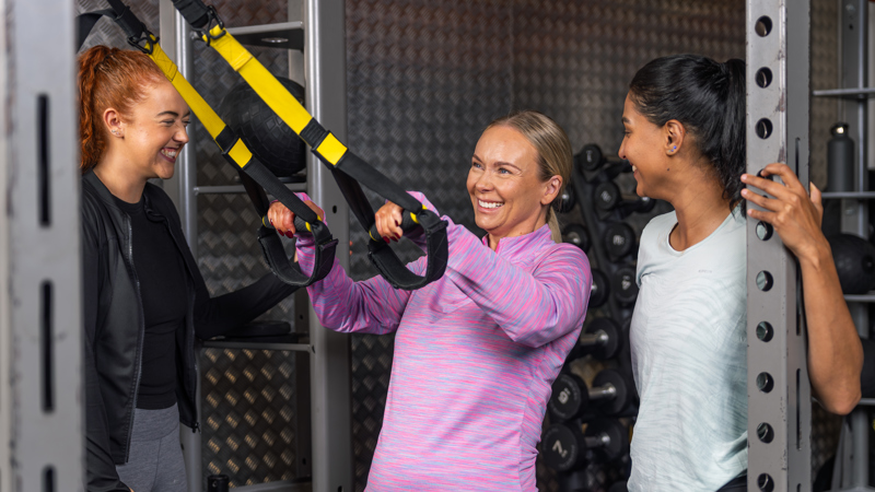 Three women smile and talk in a gym as the woman in the centre holds onto yellow suspension trainer straps.