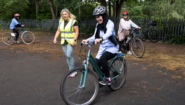 A young lady with a big smile on her face learning to ride a two wheeled bicycle with help from a coach wearing a high vis yellow vest