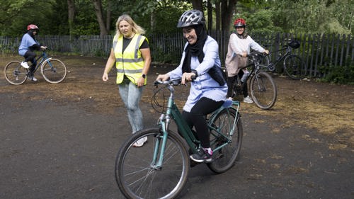 A young lady with a big smile on her face learning to ride a two wheeled bicycle with help from a coach wearing a high vis yellow vest