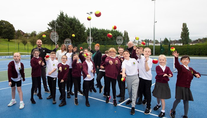 School children at Garrowhill Park tennis courts throwing tennis balls towards the camera with adults holding rackets up behind them.