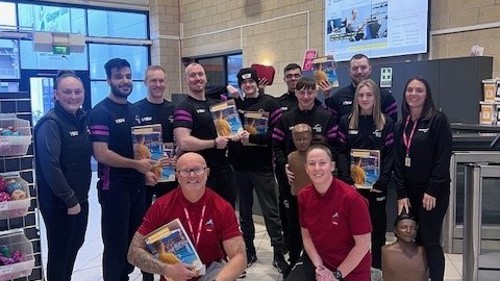 A group of 11 people gathered for a photo in a leisure centre reception area. Some of them are holding certificates and two are holding CPR dummies