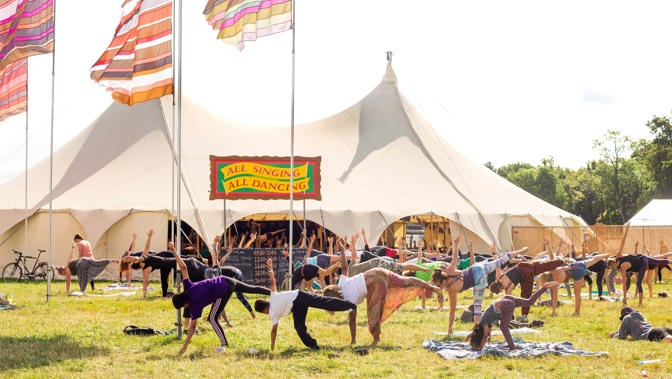 A group of people doing yoga at a music festival on a sunny day in front of a big white tent.