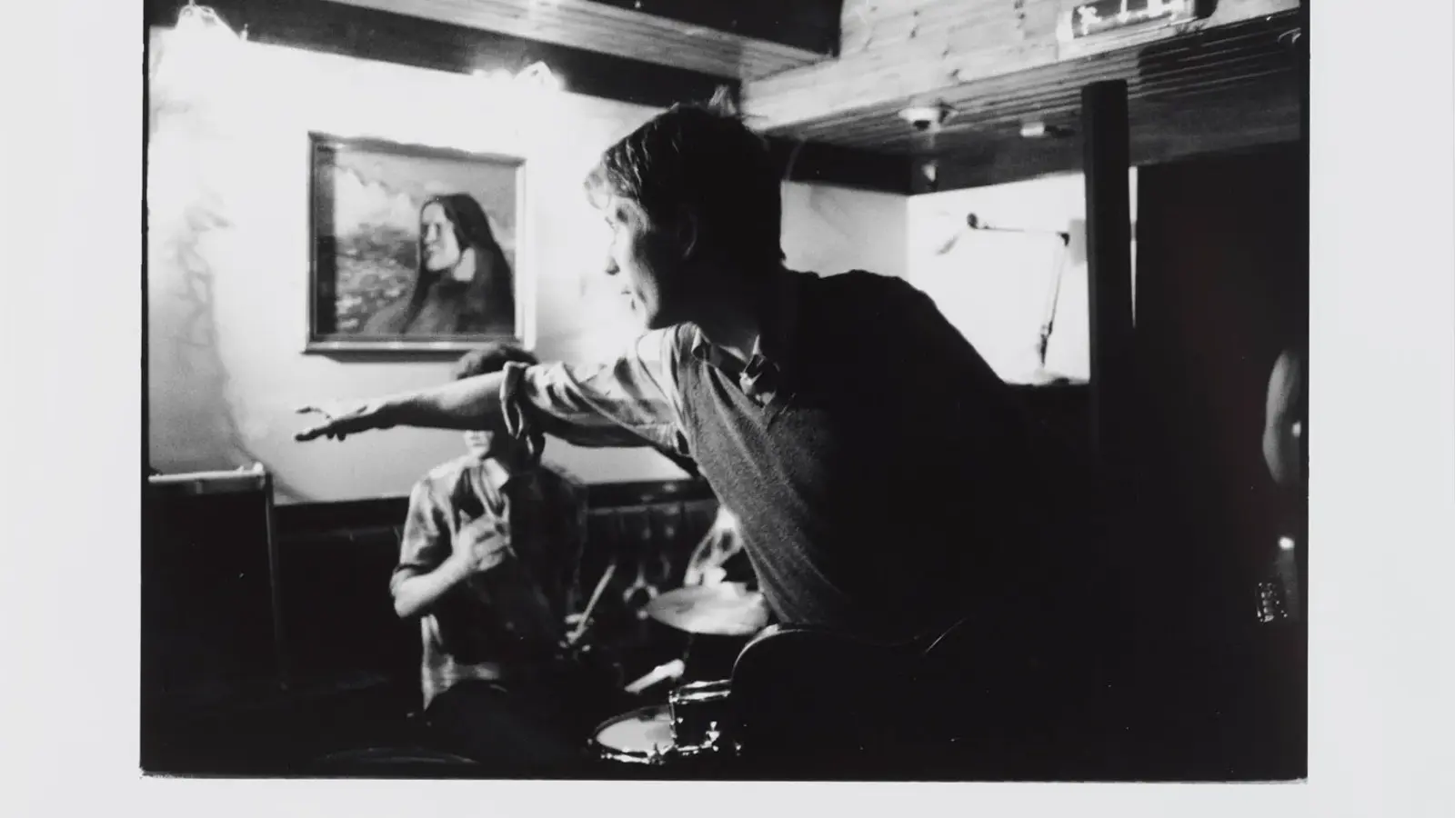A black‑and‑white photo shows a small indoor music performance. In the foreground, a guitarist extends an arm outward while playing, their gesture caught mid‑motion. Behind them, a drummer sits at a drum kit, drumsticks in hand. The room feels intimate, with a low ceiling, visible lighting fixtures, a microphone stand, and a framed portrait on the wall. The overall atmosphere suggests a candid moment during a live set in a cosy venue