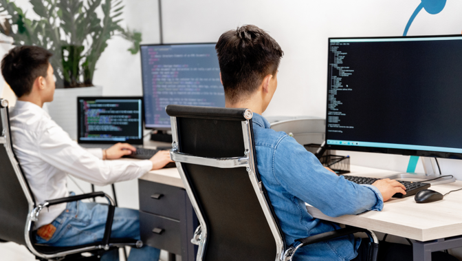 Two students with their backs to the camera sitting in an office chair facing a PC, the screen on the PC showing computer code.