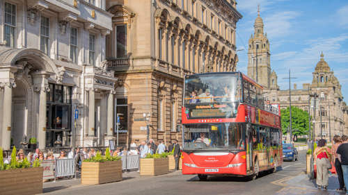  A red double decker bus is driving on a road, people are sitting at tables enjoying food and drink, there is sandstone buildings and blue skies behind the bus