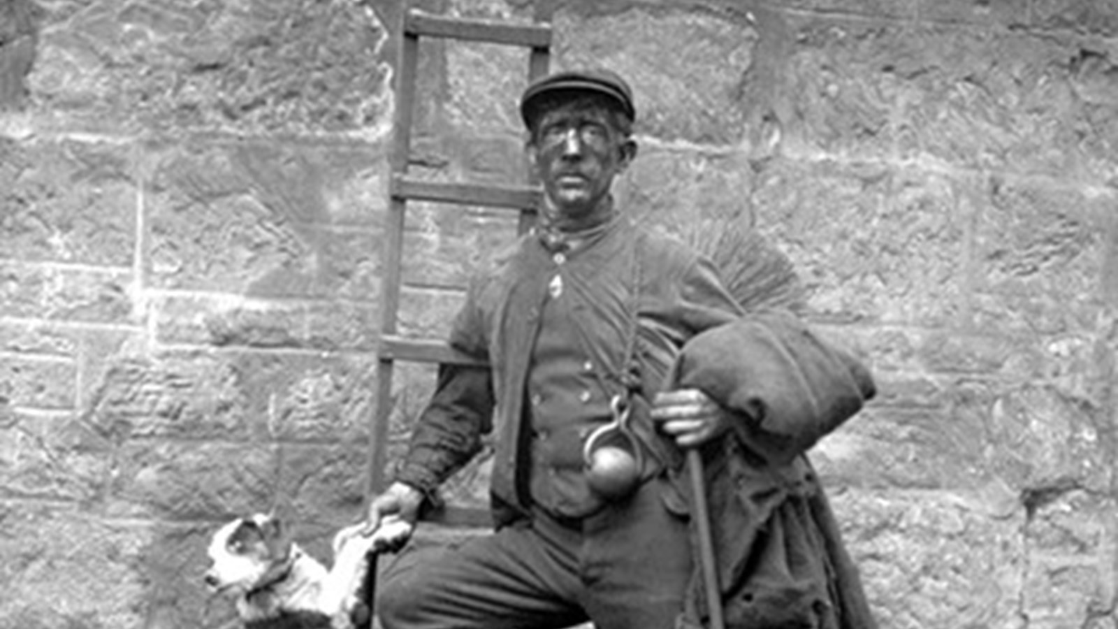 A black and white photo of a Glasgow sweep worker, with a face dirty with soot, leaning on a ladder, their jacket hanging over their left arm and a black and white dog standing on back paws with front paws in it's owners' hands.