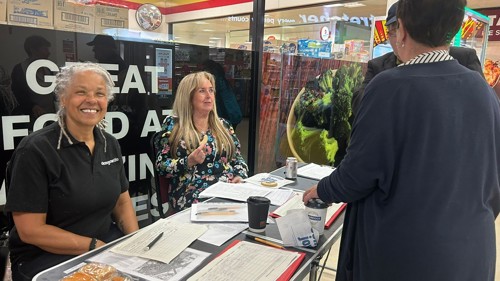 Two people are sitting behind an information table in a shopping centre, talking to members of the public