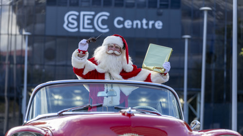 Santa holding a gold-wrapped present leaning out of a classic red American convertible from the 1950s