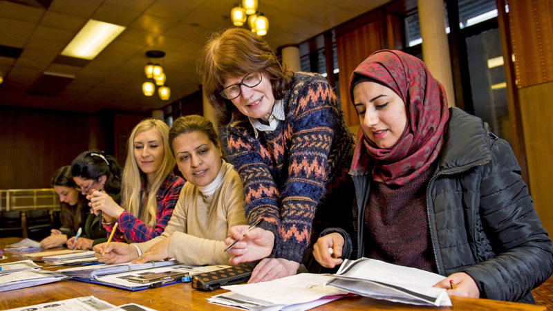 Five women sitting at a desk in a library reading and writing while listening to a tutor who is speaking to them from over their shoulders