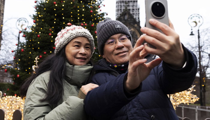 Two people taking a selfie in front of a large outdoor Christmas tree adorned with colourful lights, with a historic building and festive decorations visible in the background.
