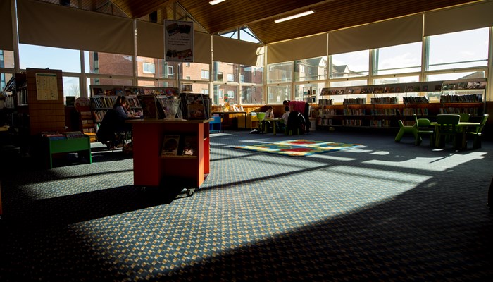 A wide view of the spacious children's area. The photograph looks like it's been taken in late afternoon. It shows sunshine outside and the inside of the library looks shadowy and atmospheric. The sun is shining on the blue checked carpet and you can see shadows and outlines of the bookshelves, tables and chairs and PC area near the windows