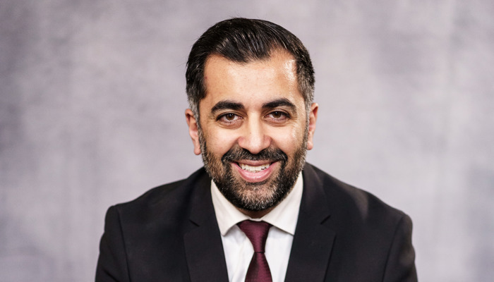 A mean wearing a dark suit and tie and white shirt is sitting down