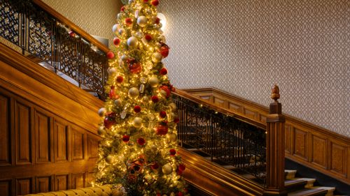 A large, beautifully decorated Christmas tree stands at the foot of an ornate wooden staircase in an elegant interior. The tree is covered with warm white lights and red and gold ornaments. The staircase features dark wrought-iron railings and rich wood panelling, with patterned wallpaper in the background”