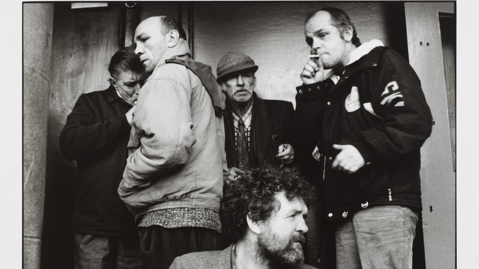 A black‑and‑white street photograph of six adults standing close together near a building entrance. They wear heavy jackets in what appears to be cold weather. One person smokes while another speaks, and the group’s candid expressions suggest an informal, everyday moment in an urban setting.