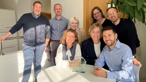 A group of eight people gathered around a glass award in a sunny open plan office. Three of them are sitting at a small round table with the rest standing behind