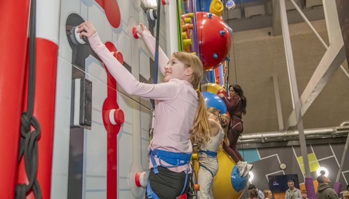 A smiling young girl with a pink zipper and blue harness on climbing up a climbing wall.