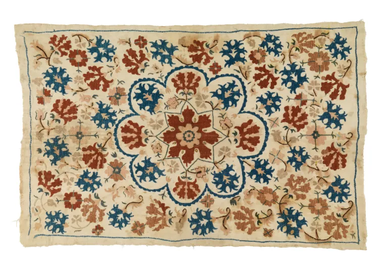 an image of an embroidered rug with red and blue patterns on a cream base.