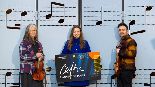 A group of three people wearing winter coats standing in front of a set of doors with musical notes printed on them. One of them is holding a violin
