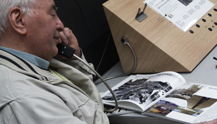 An elderly community member, engaged with an archival art exhibition. They are listening to sound work through a telephone receiver and smiling. 
