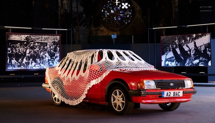 A red car draped in a vast net curtain sits in front of two large vide screens 