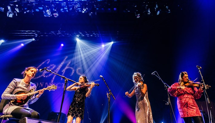Four people onstage playing instruments. One is seated playing the mandolin and the other three are playing fiddles. All are wearing sequinned dresses.
