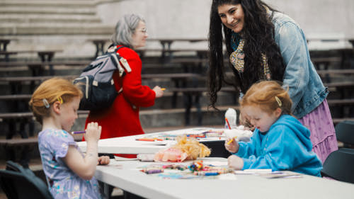 Two young girls sit at a table doing arts and crafts while a woman leans over, smiling and engaging with them; another woman walks by in the background.