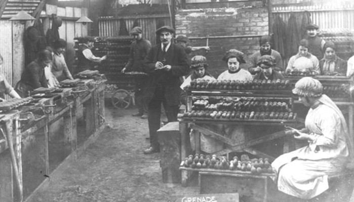 A black and white photo of several women workers in a factory creating grenades. A man in a dark suit is walking around taking notes.