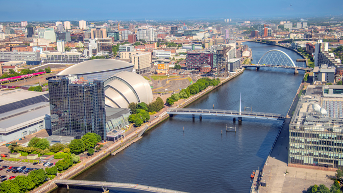 Aerial view of a cityscape with a wide river running through the centre, crossed by multiple bridges, including a distinctive white arch bridge in the distance. In the foreground sits a large modern concert venue with a curved metallic roof beside a glass-fronted building. Streets, offices, and residential buildings extend across both sides of the river under a clear blue sky.