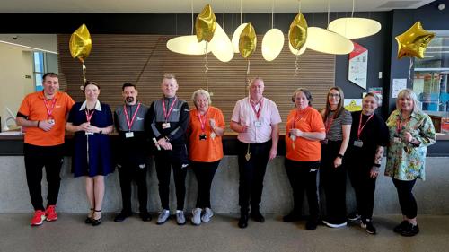 A group of ten people standing in front of a reception area in a sports centre. They are posing for a photograph with gold star-shaped helium balloons