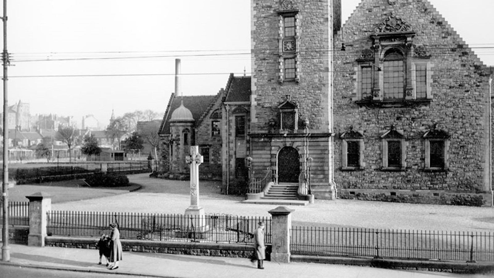 A black and white photo of Pollokshaws Burgh Hall that has a large cross headstone in the grounds with people walking by on the pavement.