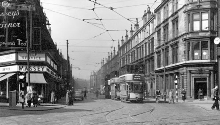 A black and white photo of a street with many trams driving along the lines, tall tenement style shop buildings are on both sides with people walking on the pavement.