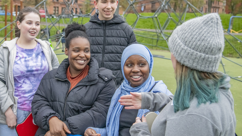4 people are looking and smiling towards a 5th person who has their back to the camera. There is a playground in the background