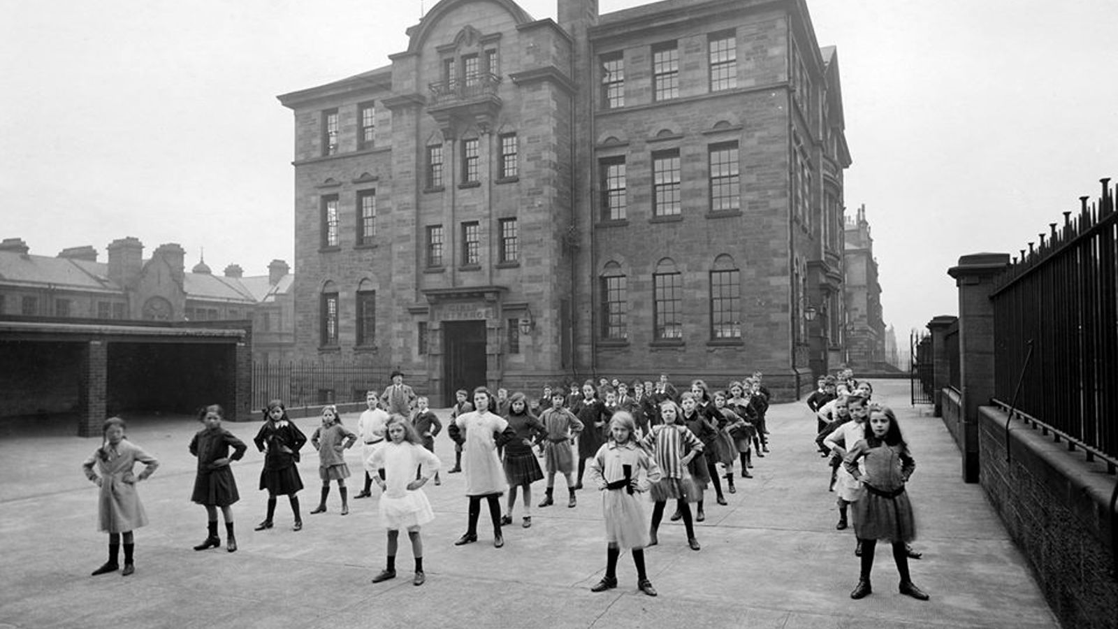 A black and white photo of a class of children standing outside of their large red sandstone school, posing in their uniforms with their hands on hips.