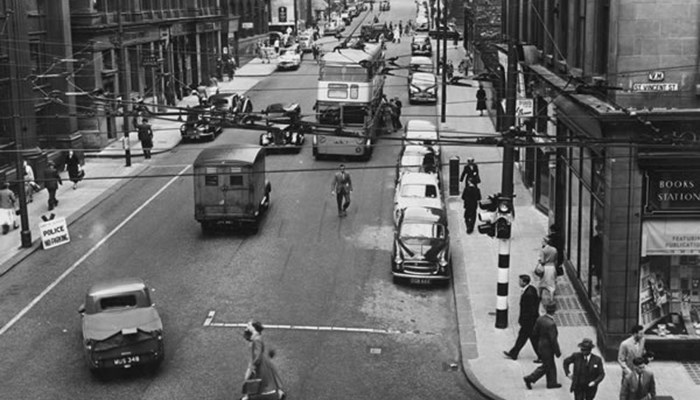 A black and white photo of a street in Glasgow with pedestrians on both the pavement and crossing the road and many cars and buses driving on the road. Both sides have tall tenement buildings.